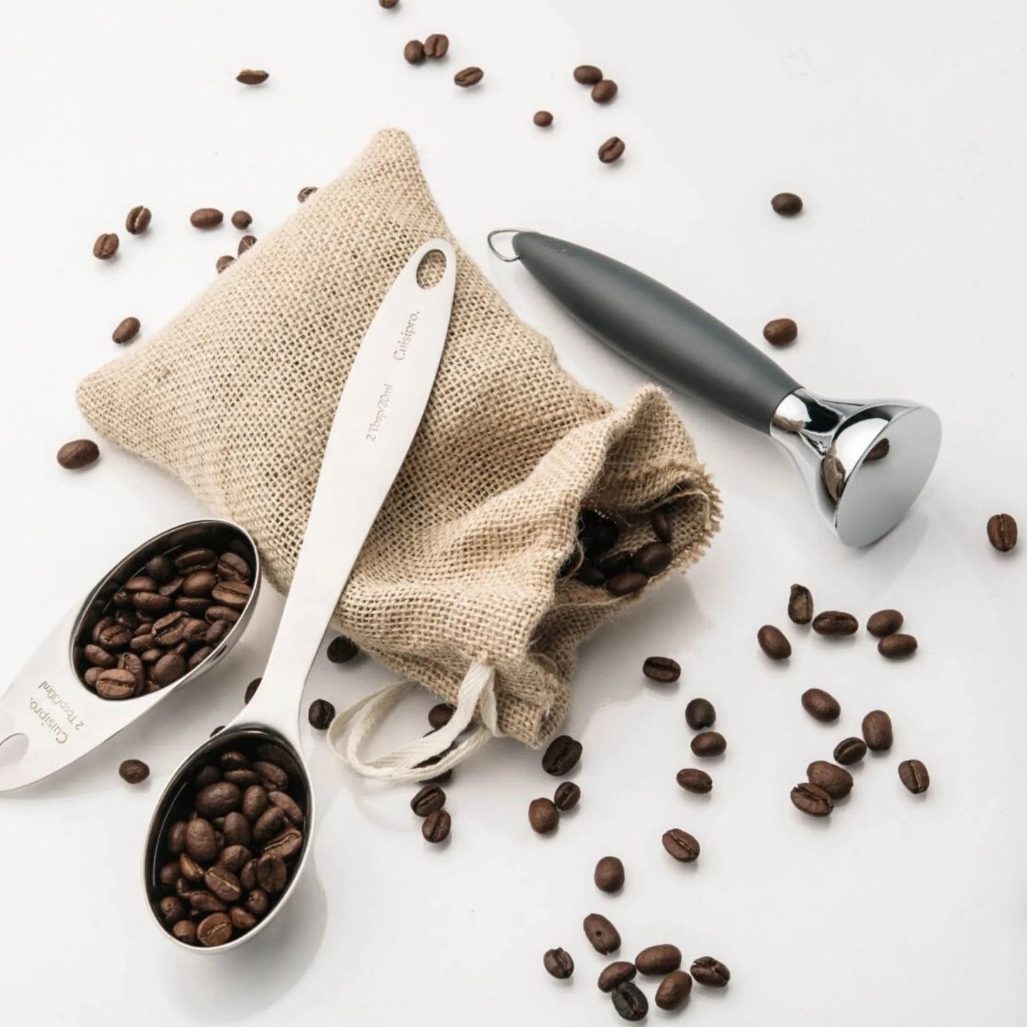 Stainless steel coffee scoop with dark handle, beside linen pouch and coffee beans