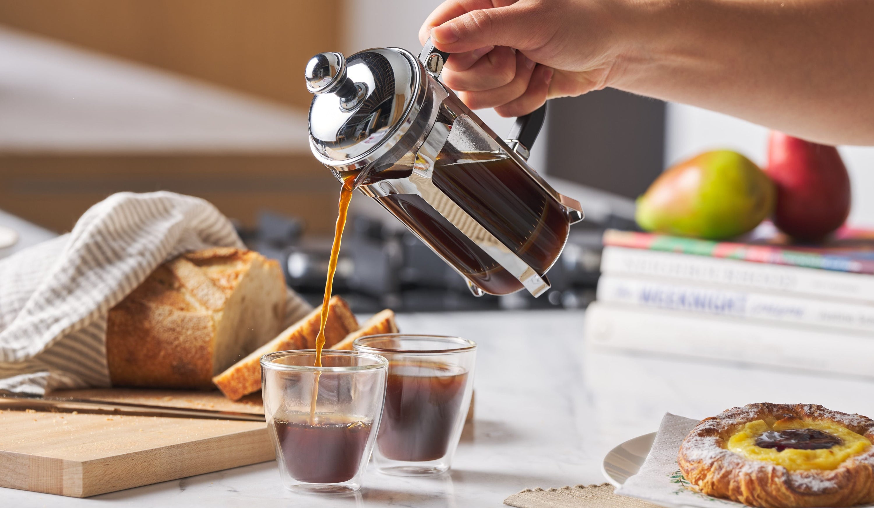 Person pouring coffee from a French press into two glasses on a kitchen counter with bread and pastries.