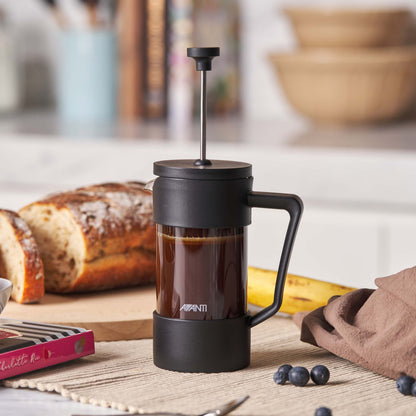 Black French press with coffee on a wooden table with bread and bananas in the background