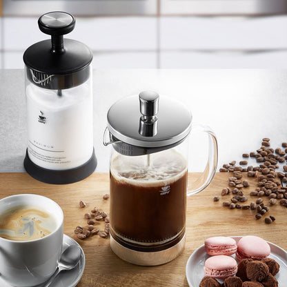 Glass coffee press with chrome plunger on wooden table, coffee beans and macaron cups nearby