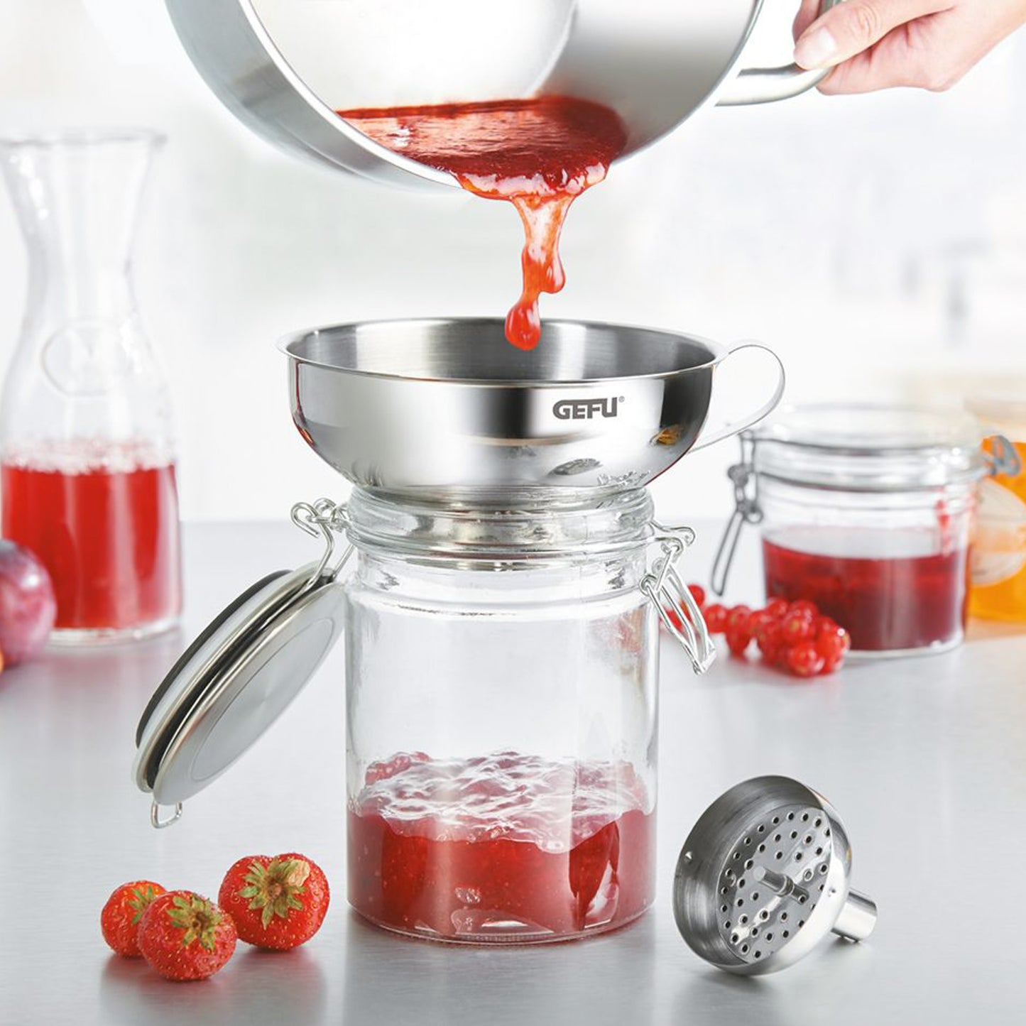 Stainless steel bottling funnel pouring into a jar, with perforated insert nearby