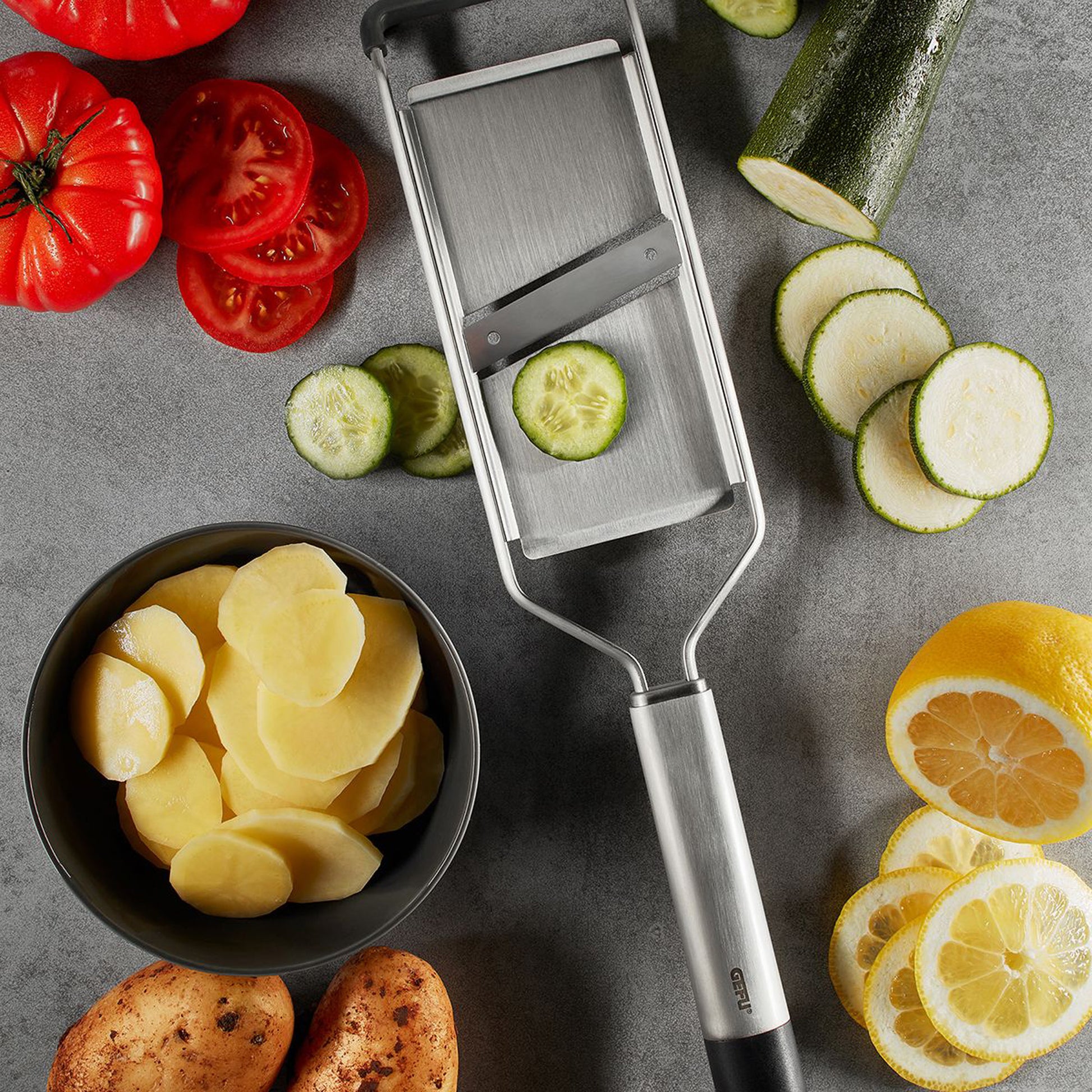 Vegetable and potato slicer with metal blade, beside sliced cucumber, potatoes, and citrus