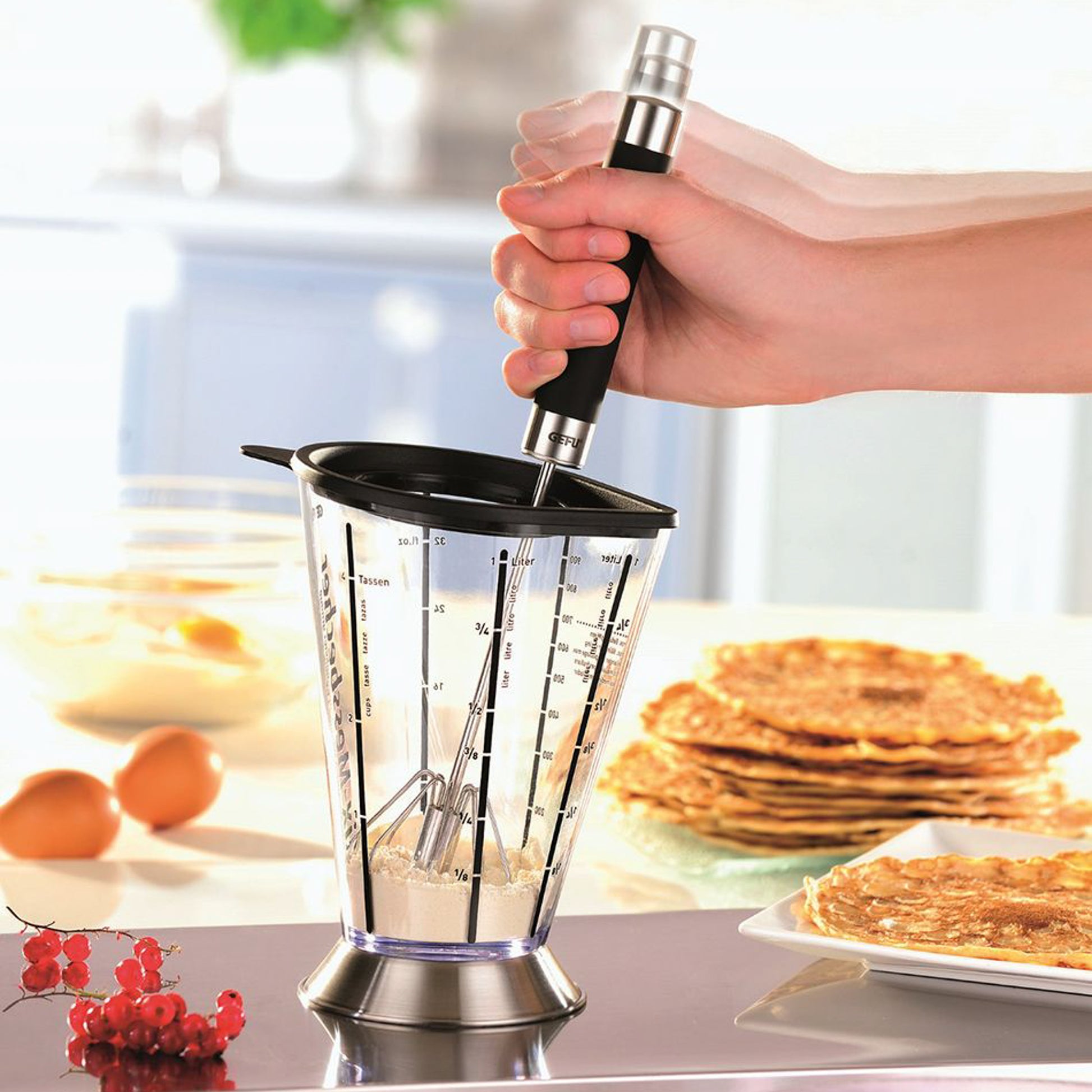 Hand holding all-purpose whisk with black handle over mixing cup and batter