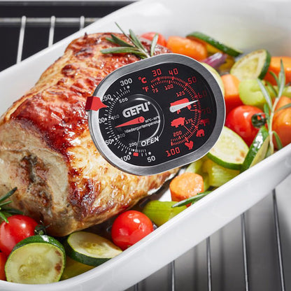 Oven thermometer on meat and vegetables in baking tray, round black dial with red markings