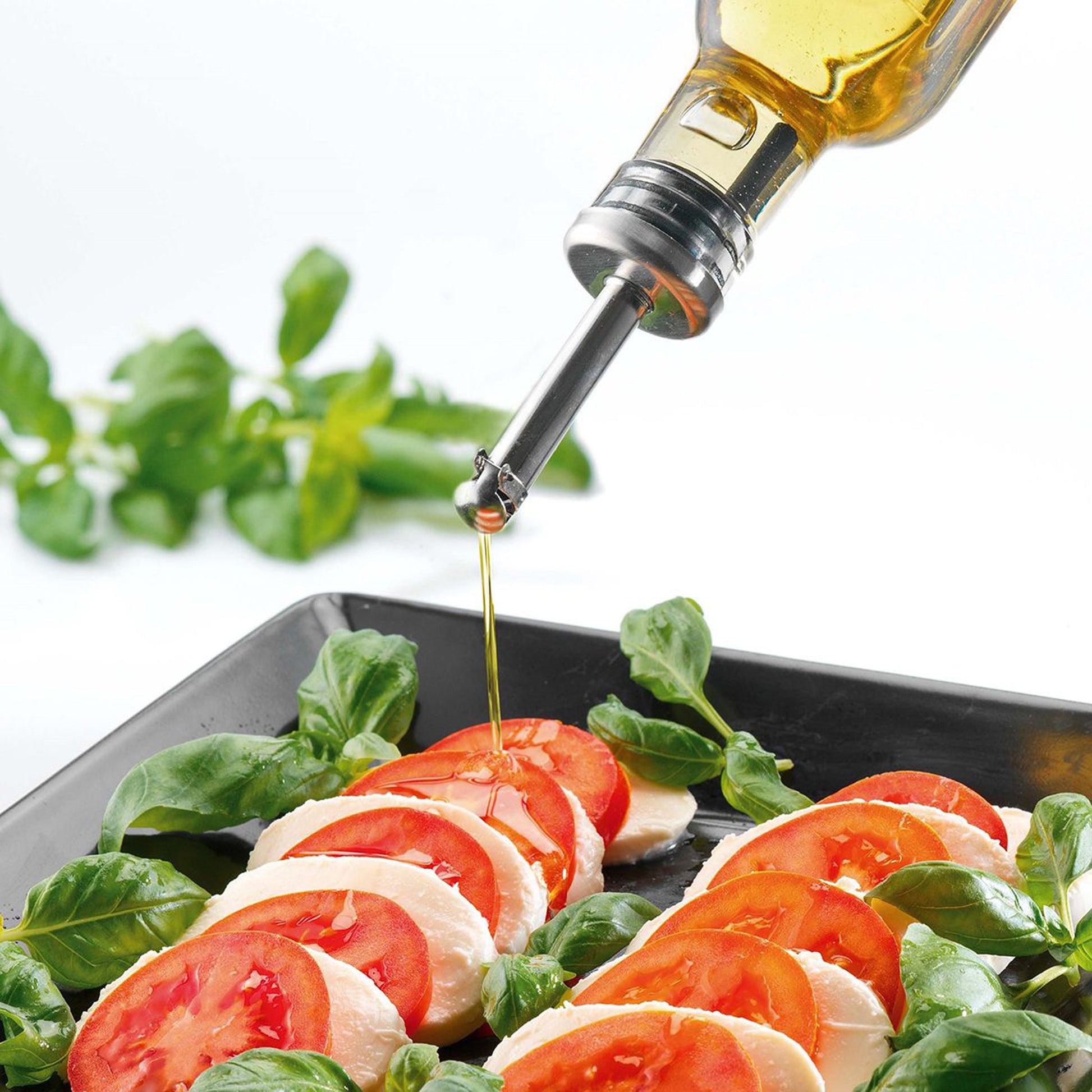 Pourer spout pouring oil onto tomato slices and basil leaves in a tray