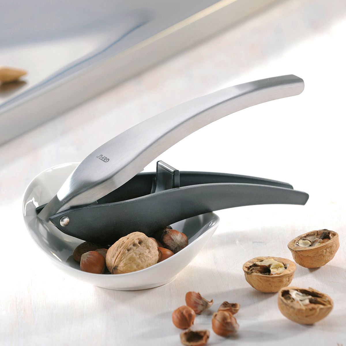 Metal nutcracker with curved silver lever and dark handle, with nuts and shells nearby