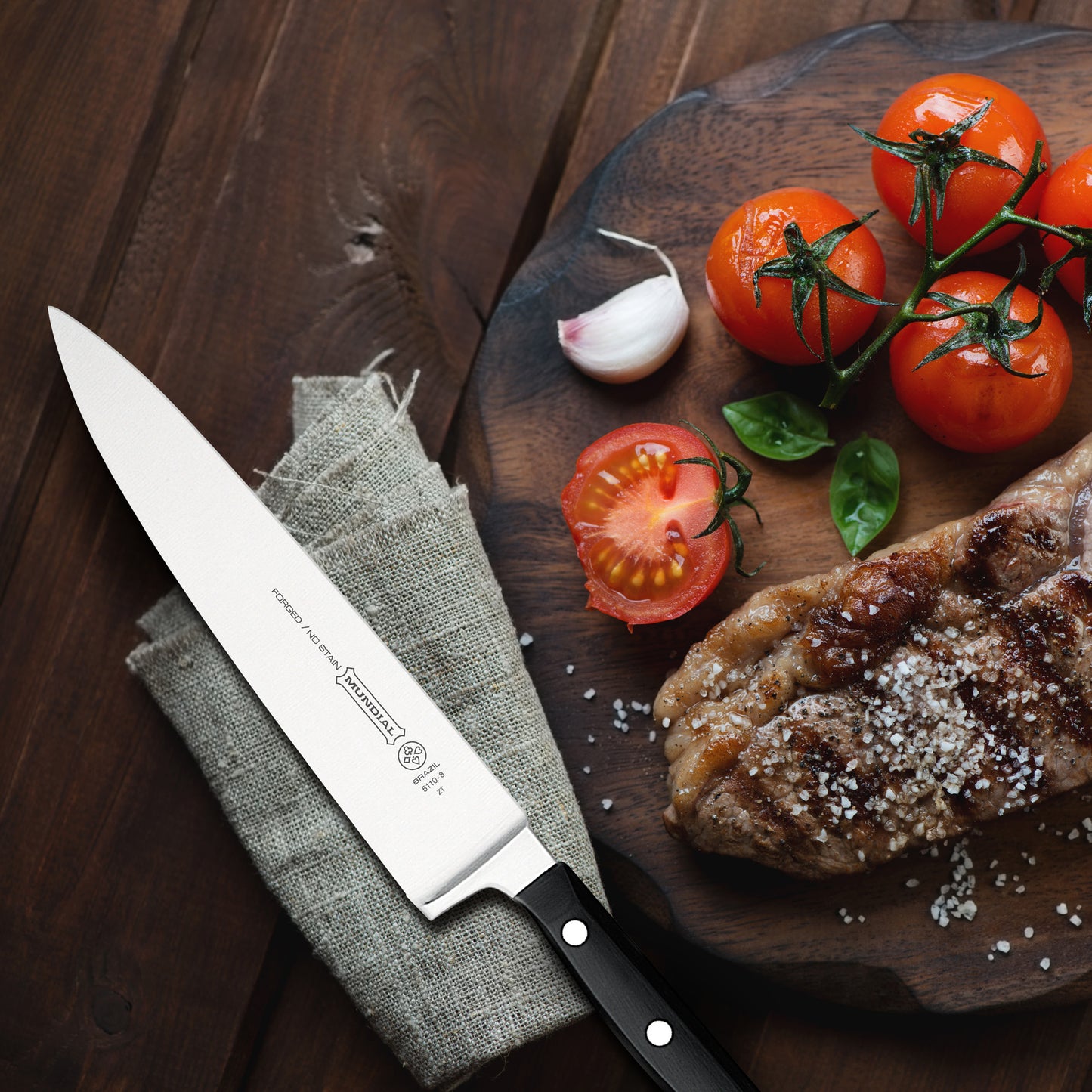Chef's knife on gray fabric with black handle, beside tomatoes and herbs