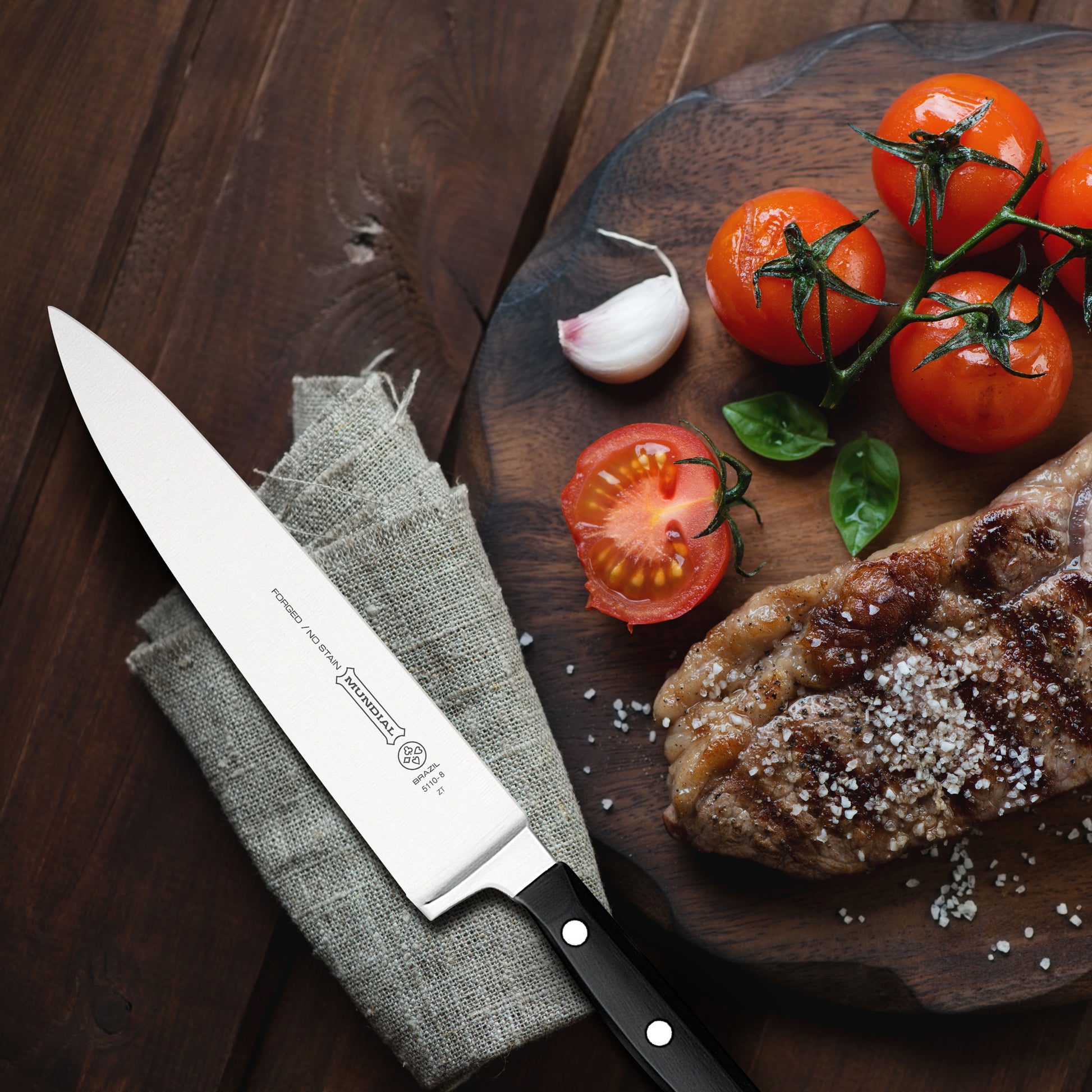 Chef's knife on gray fabric with black handle, beside tomatoes and herbs