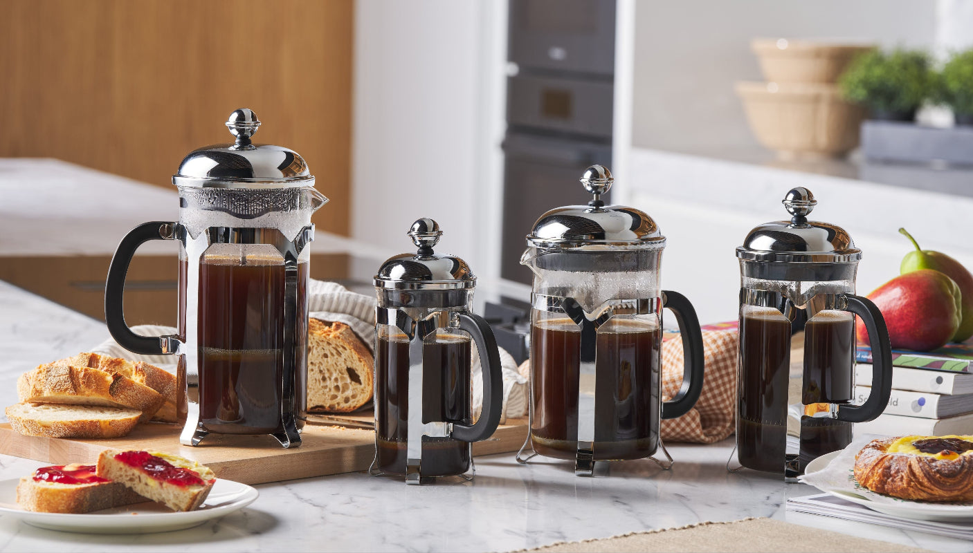 Set of four French press coffee makers on a kitchen counter with pastries and fruit.