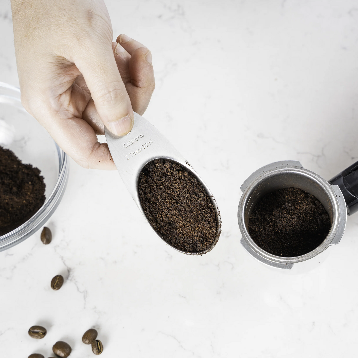 Hand holding Cuisipro coffee scoop with dark grounds over jar and bowl