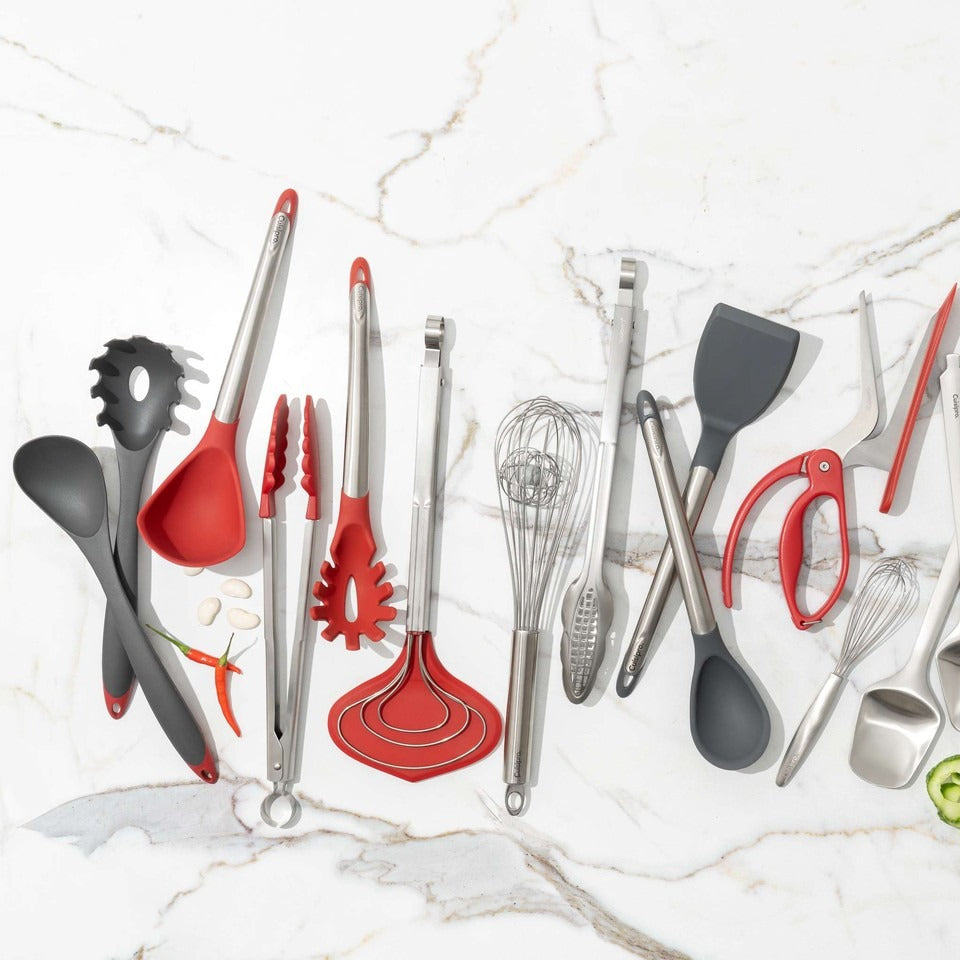 A set of kitchen utensils including spatulas, whisks, tongs, and spoons in red, black, gray, and silver, arranged on a white marble background.
