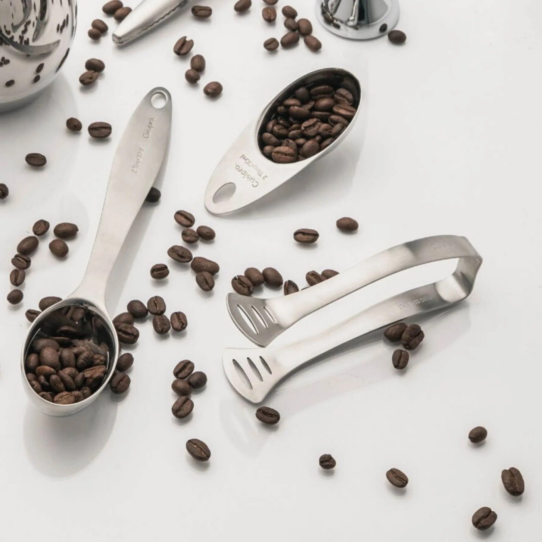 Pair of stainless steel tea bag tongs lying on a countertop with coffee beans